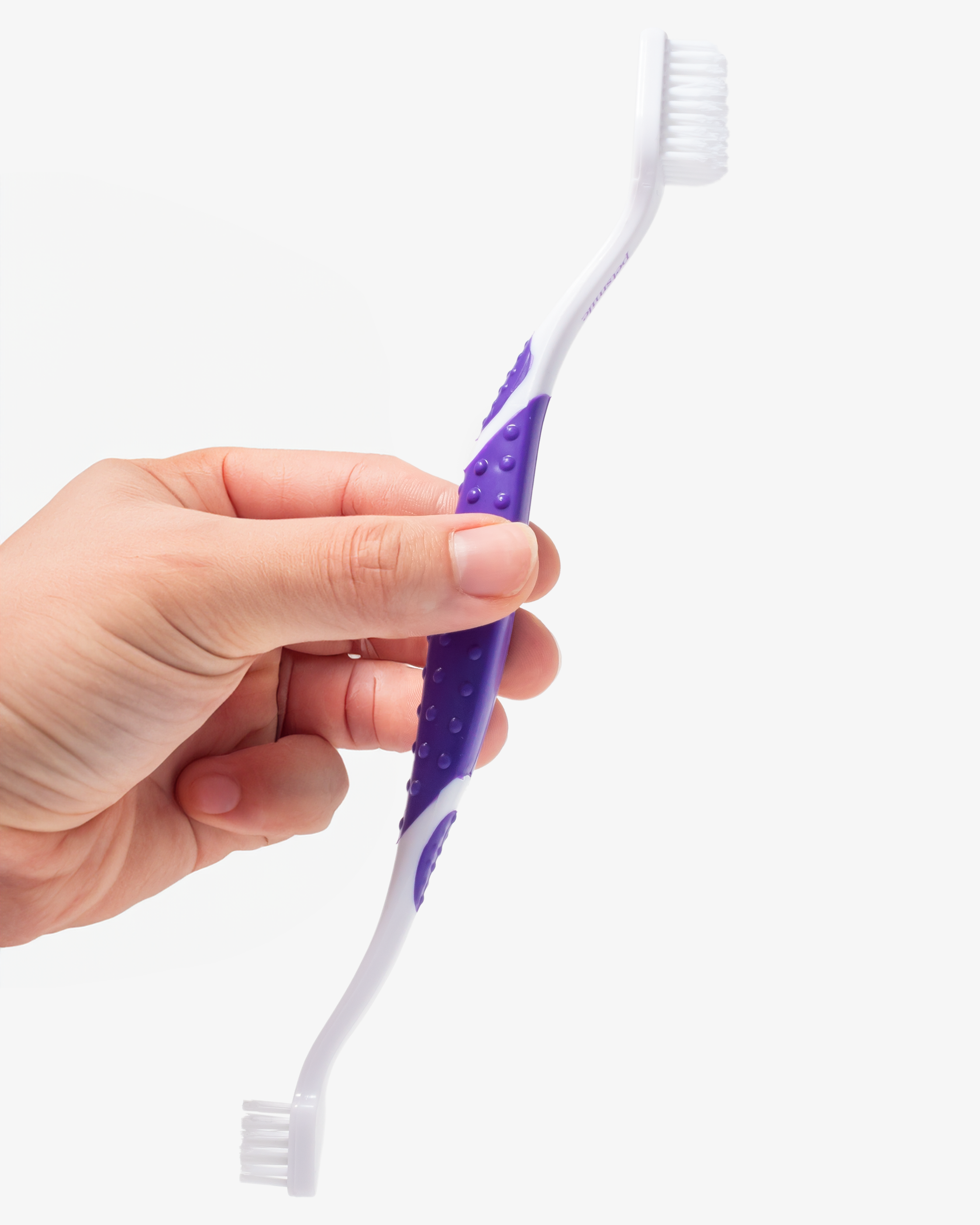 A hand holds the petsmile Professional Pet Toothbrush, featuring a purple rubber grip and white bristles, perfect for dental care with Professional Toothpaste Rotisserie Chicken Small, set against a plain white background.
